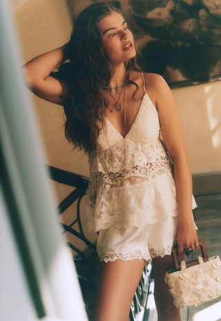 a-woman-wearing-a-cream-lace-camisole-top-and-matching-lace-shorts-holding-a-small-cream-floral-handbag-posed-in-a-warm-toned-indoor-setting