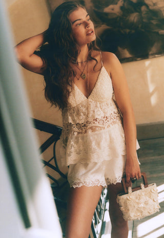 a-woman-wearing-a-white-lace-camisole-top-and-matching-lace-shorts-holding-a-small-white-floral-handbag-in-a-sunlit-indoor-setting